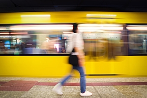 Im Vordergrund ist eine laufende Frau und im Hintergrund eine gelbe Bahn, die an ihr vorbeifährt.