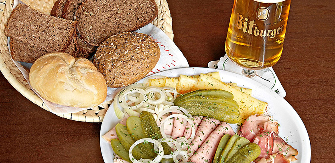 Vesperteller mit Wurst, Käse und Gurken neben einem Brotkorb und ein Glas Bier