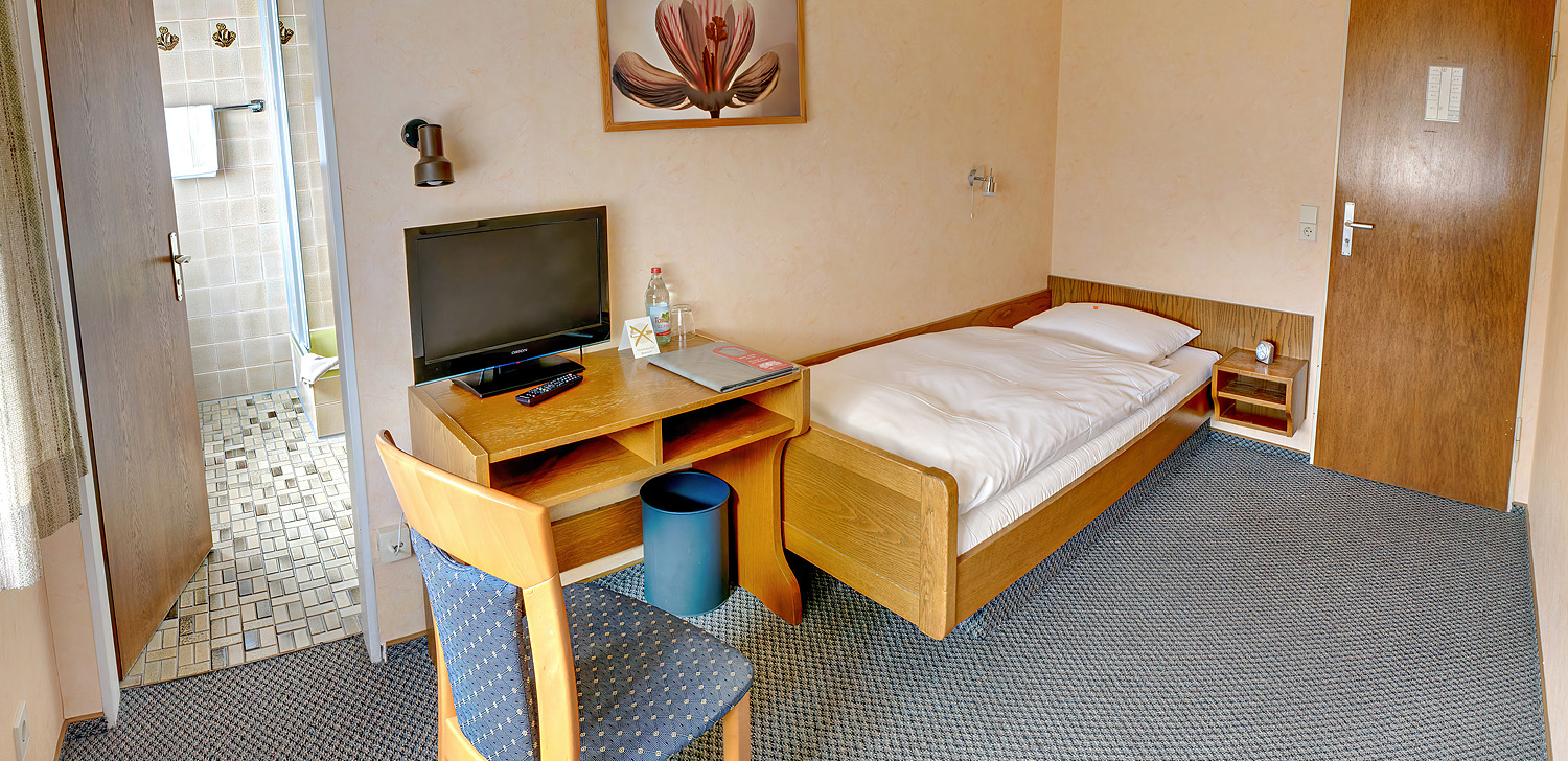 Helles Hotelzimmer mit einem Einzelbett an einer Wand, einem Tisch mit Fernseher darauf und Stuhl.